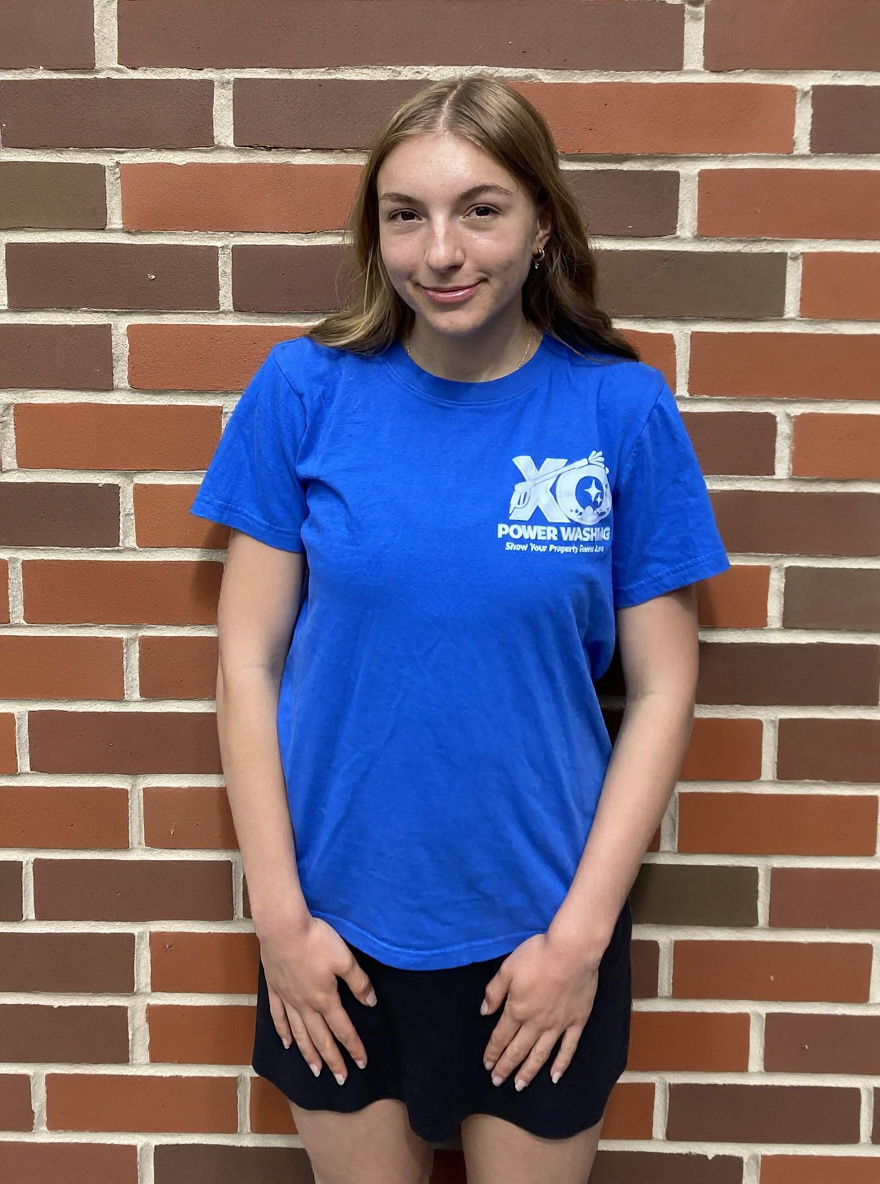 Grace, customer service lead at XO Power Washing, smiling in a branded blue shirt in front of a brick wall
